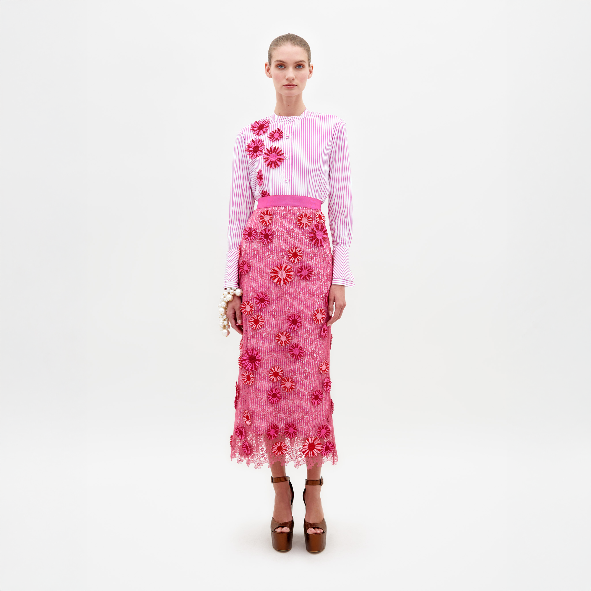 Against a plain white background, a person dons a striped pink and white top with 3D floral appliqué, paired with the Uma Skirt by SVT DESIGNS, which showcases floral patterns and a scalloped lace hem. Brown high heels complete the look, and their hair is styled up.