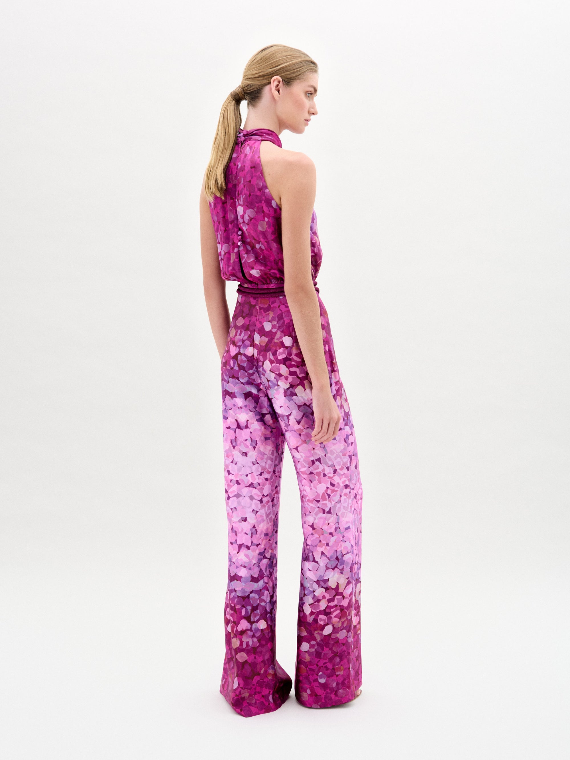 A woman stands before a simple white backdrop, styled in the Siv Jumpsuit by SVT DESIGNS—a sleeveless floral piece in pink and purple, made from luxurious Italian fabric. Her hair is tied back in a ponytail and she turns slightly to the side.