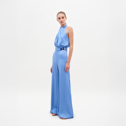 A person models the Siv Jumpsuit by SVT DESIGNS against a white backdrop. The blue hammered satin fabric features a halter-neck, cinched waist with belt, and wide-legged trousers, complemented by black sandals.