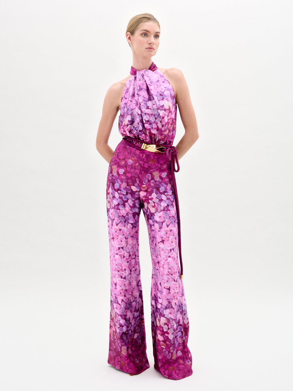 A person poses against a white background wearing the Siv Jumpsuit by SVT DESIGNS—a sleeveless halter-neck piece made from Italian fabric with a pink and purple abstract print, wide legs, and a thin waist-cinching belt.