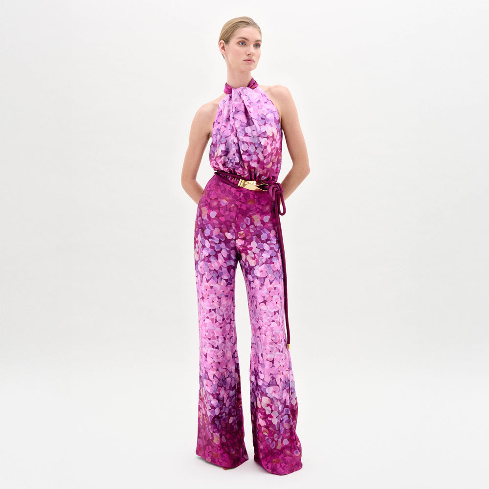 A person poses against a white background in the Siv Jumpsuit by SVT DESIGNS, featuring an abstract print in pink and purple, a halter-neck, flared legs, and a matching belt, all crafted from premium Italian fabric.