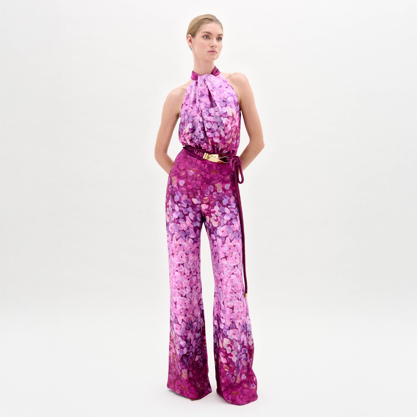 A person poses against a white background in the Siv Jumpsuit by SVT DESIGNS, featuring an abstract print in pink and purple, a halter-neck, flared legs, and a matching belt, all crafted from premium Italian fabric.