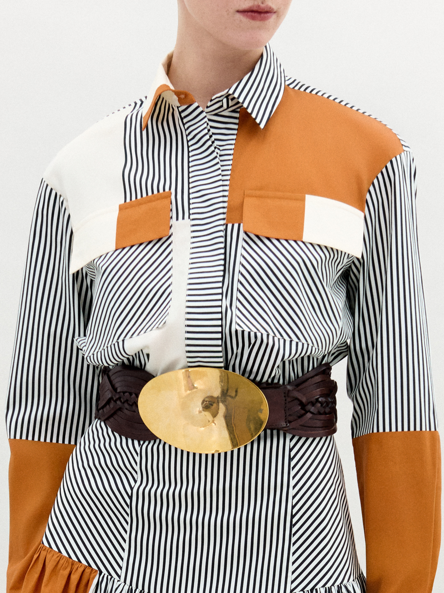 A person wearing the Soleil Blouse—long-sleeved and striped with orange and white color blocks in 100% Italian cotton—paired with a large gold belt, stands against a plain light background.