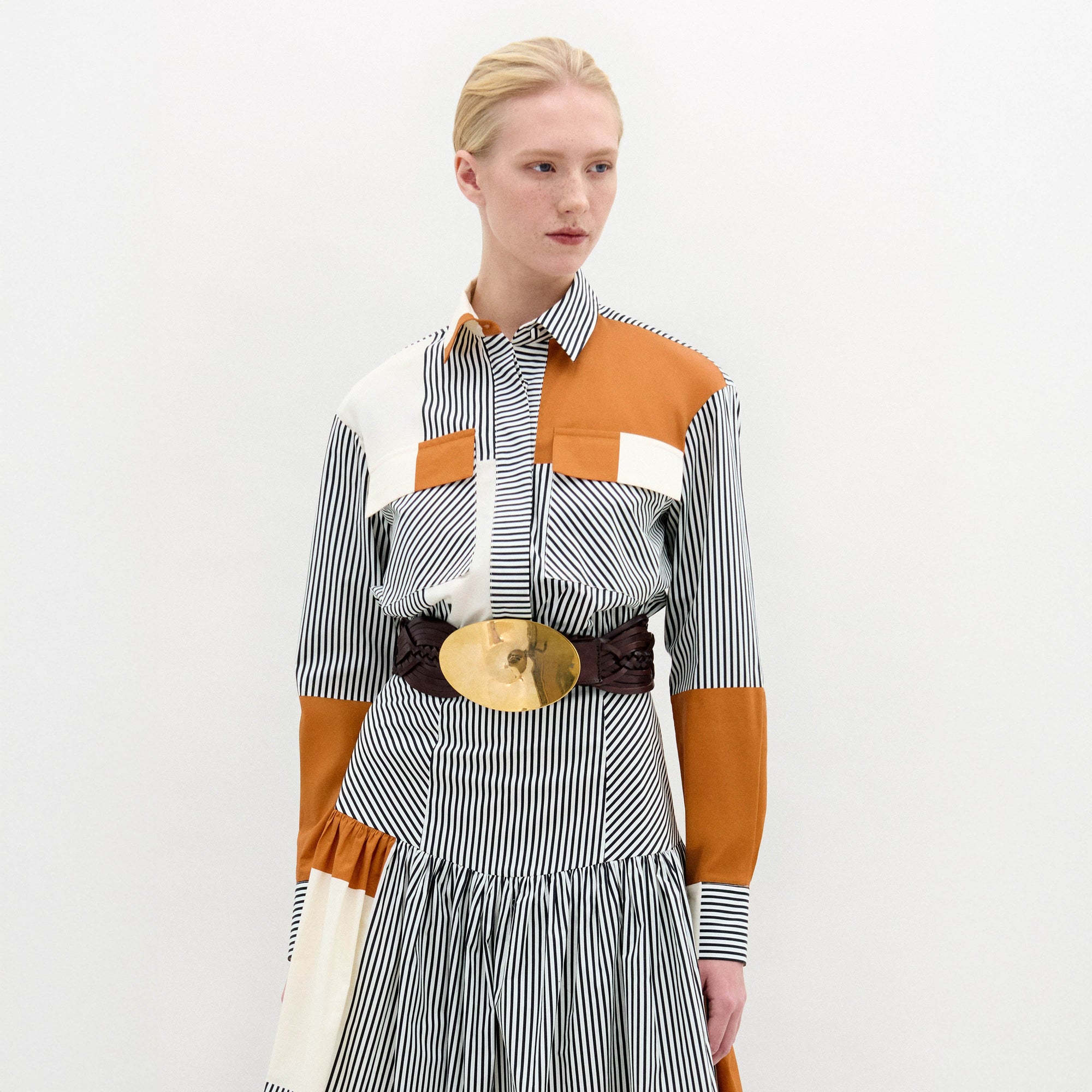 A person wearing the Soleil Blouse—long-sleeved and striped with orange and white color blocks in 100% Italian cotton—paired with a large gold belt, stands against a plain light background.