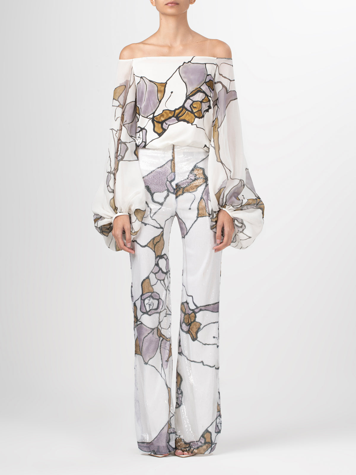 A person is wearing a Silvia Tcherassi US Avellino Pant set, featuring an off-the-shoulder sheer top with billowing sleeves and matching high-waisted wide-leg pants. The fabric showcases a mosaic-style pattern in purple, beige, and gray on a white background for an abstract print flair.