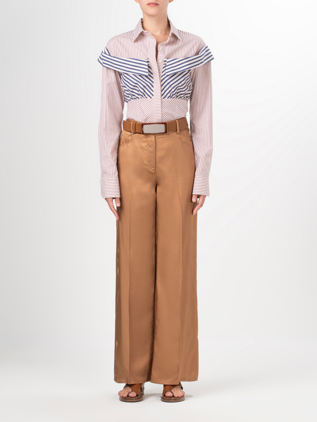 Wearing the Emine Pant by Silvia Tcherassi US, a person pairs tan, high-waisted viscose pants with a striped button-up shirt and rolled sleeves. The look is completed with sandals and a brown belt against a plain white backdrop, their face unseen.