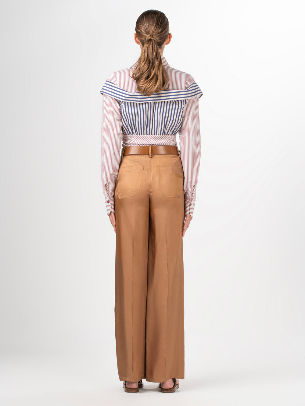 A person with long hair tied back faces away, wearing a striped off-the-shoulder blouse and the Emine Pant by Silvia Tcherassi US. These high-waisted, wide-leg brown trousers are crafted from 100% Viscose. A braided belt adds charm against the plain white backdrop.