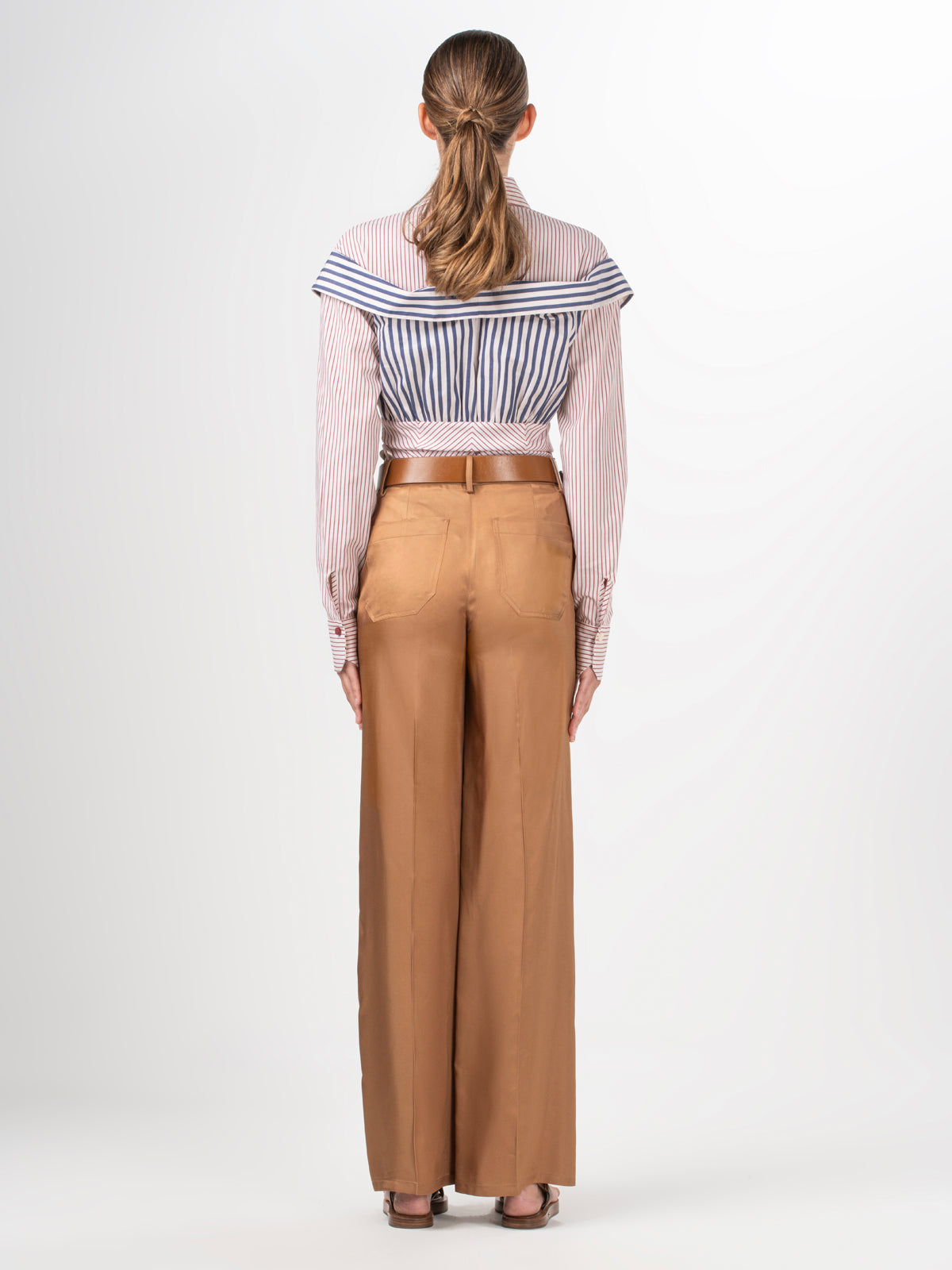 A person with long hair tied back faces away, wearing a striped off-the-shoulder blouse and the Emine Pant by Silvia Tcherassi US. These high-waisted, wide-leg brown trousers are crafted from 100% Viscose. A braided belt adds charm against the plain white backdrop.
