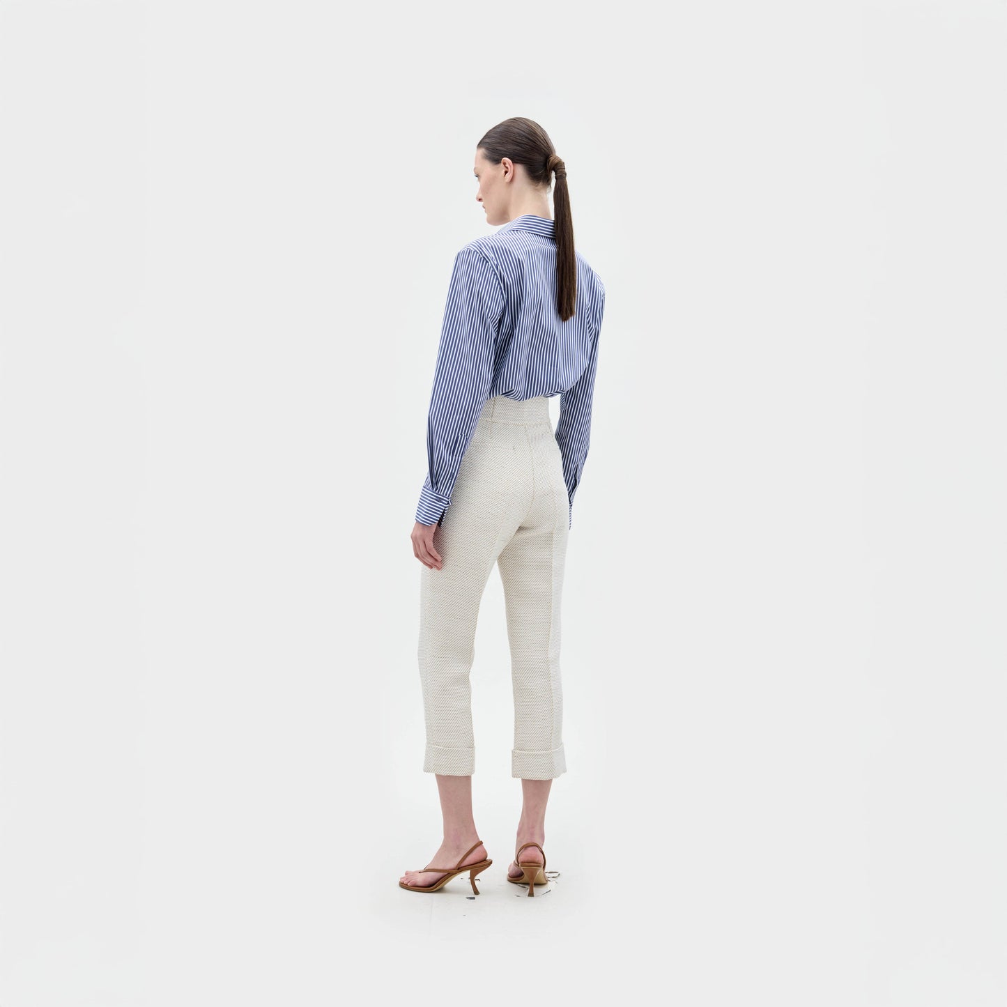A woman stands against a white background, facing away. She wears the Karole Blouse by SVT DESIGNS—100% cotton, striped, and button-down—tucked into high-waisted cream pants with brown heeled sandals. Her hair is in a low ponytail.