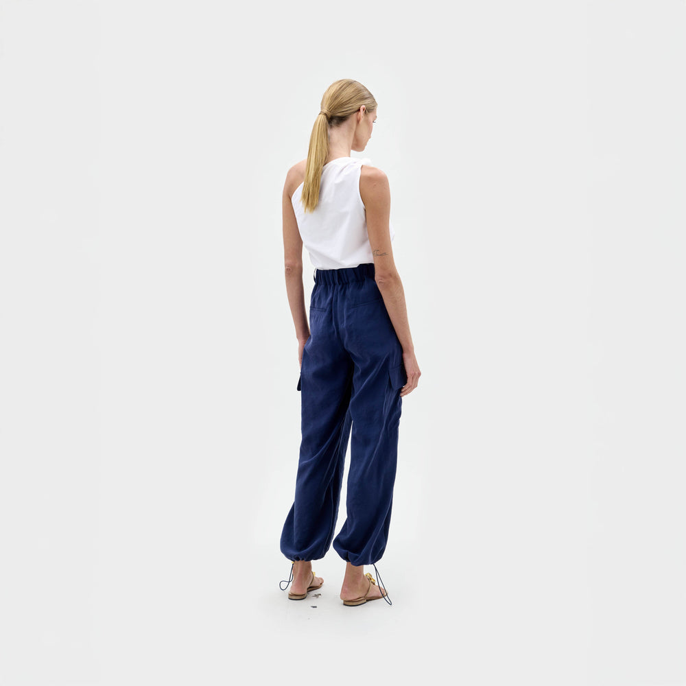A person with blonde hair in a low ponytail stands facing away from the camera, wearing SVT DESIGNS' Darnell Pant—navy blue, high waist cargo pants with elastic cuffs—a sleeveless white top, and sandals against a plain white background.
