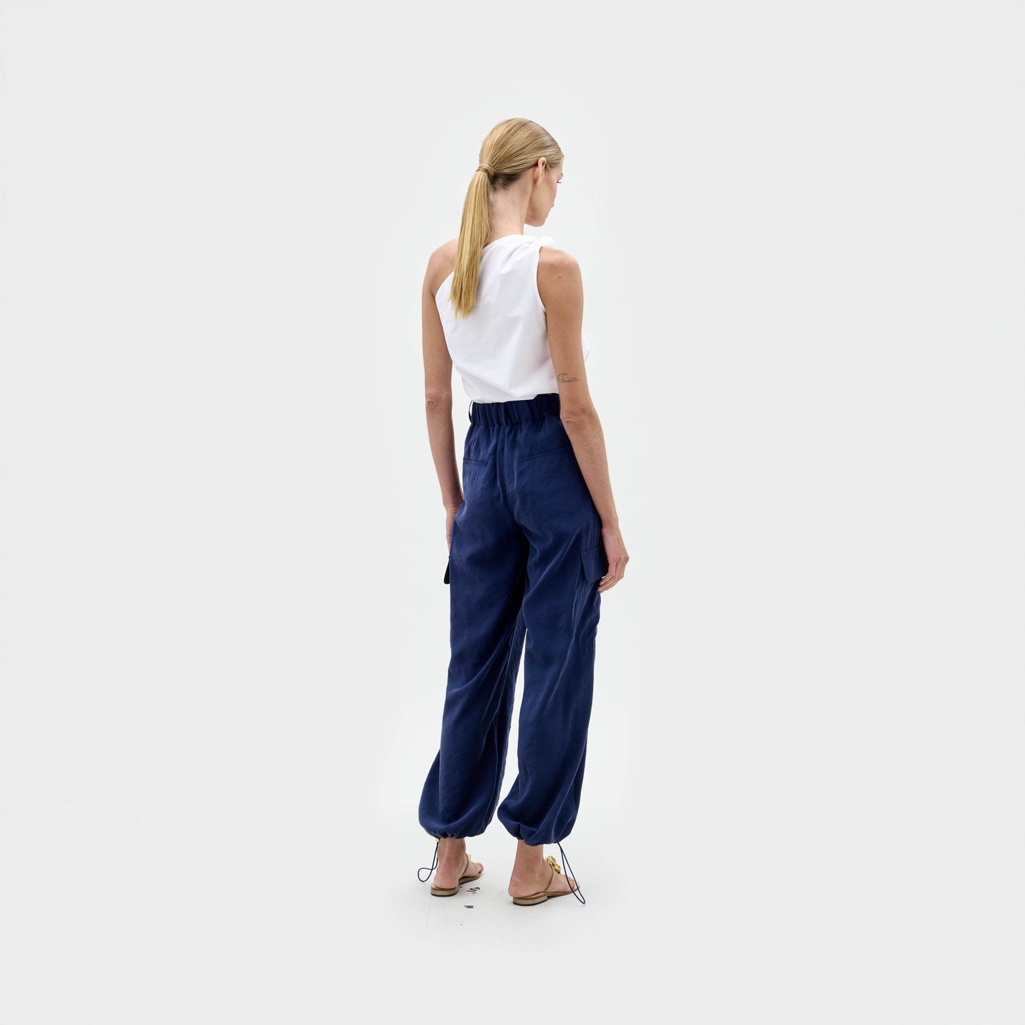 A person with blonde hair in a low ponytail stands facing away from the camera, wearing SVT DESIGNS' Darnell Pant—navy blue, high waist cargo pants with elastic cuffs—a sleeveless white top, and sandals against a plain white background.