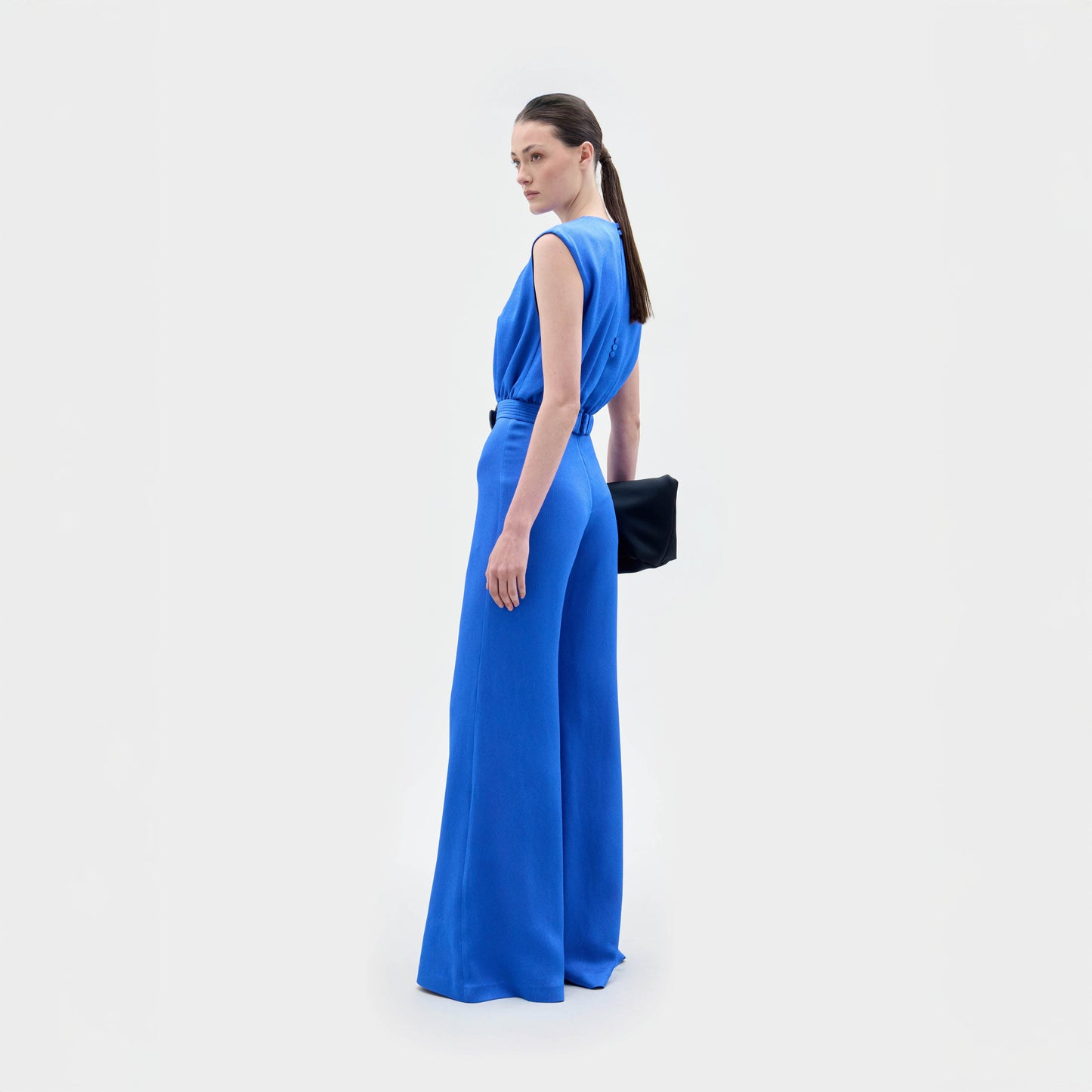 A woman poses against a white background in the SVT DESIGNS Noel Jumpsuit, a sleeveless, wide-leg piece made from Italian textile. She pairs it with a black clutch and gazes over her shoulder, turned slightly sideways.
