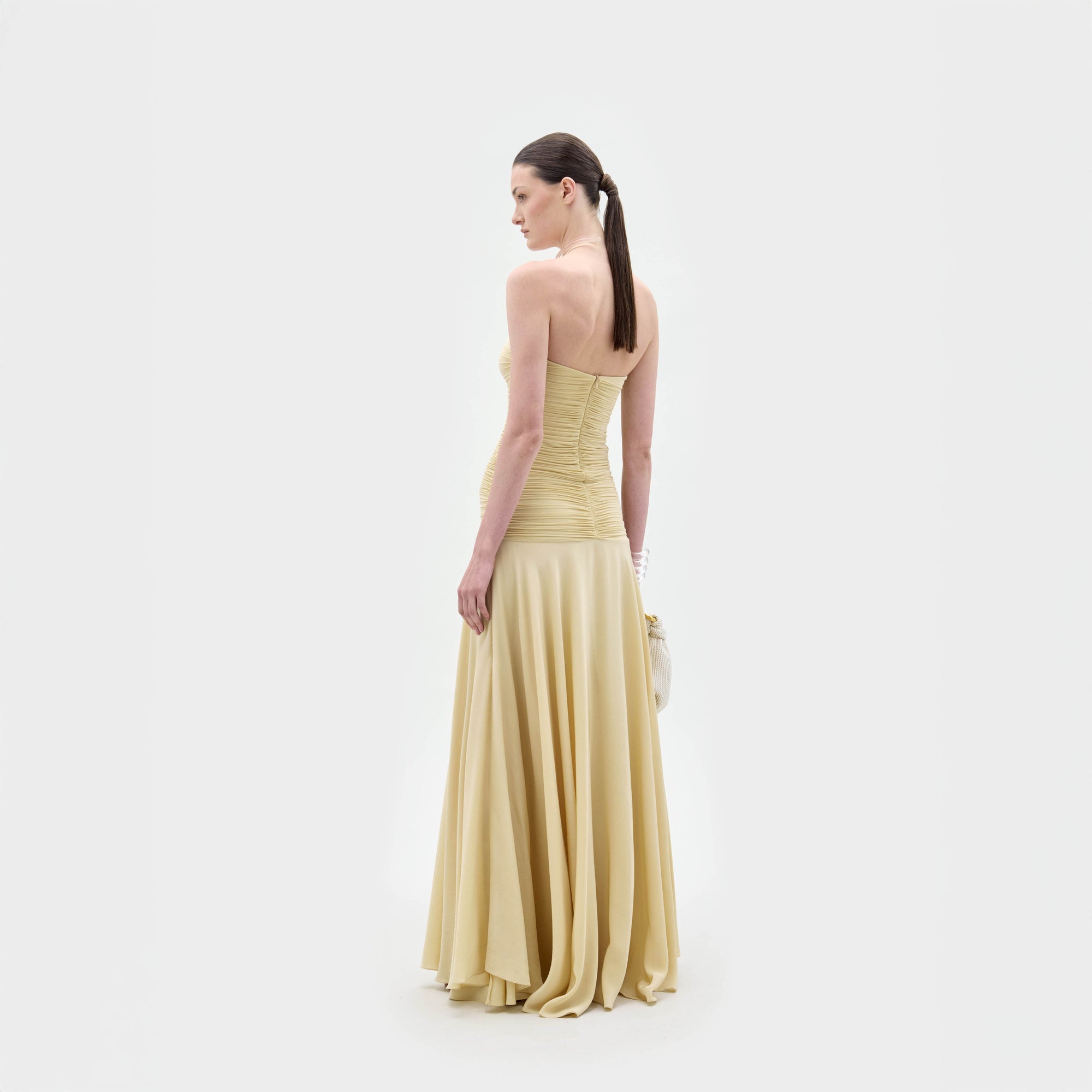 A woman with long dark hair in a ponytail faces away, wearing the SVT DESIGNS Norma Dress—a light yellow strapless maxi—while holding a small bag against a plain white background.