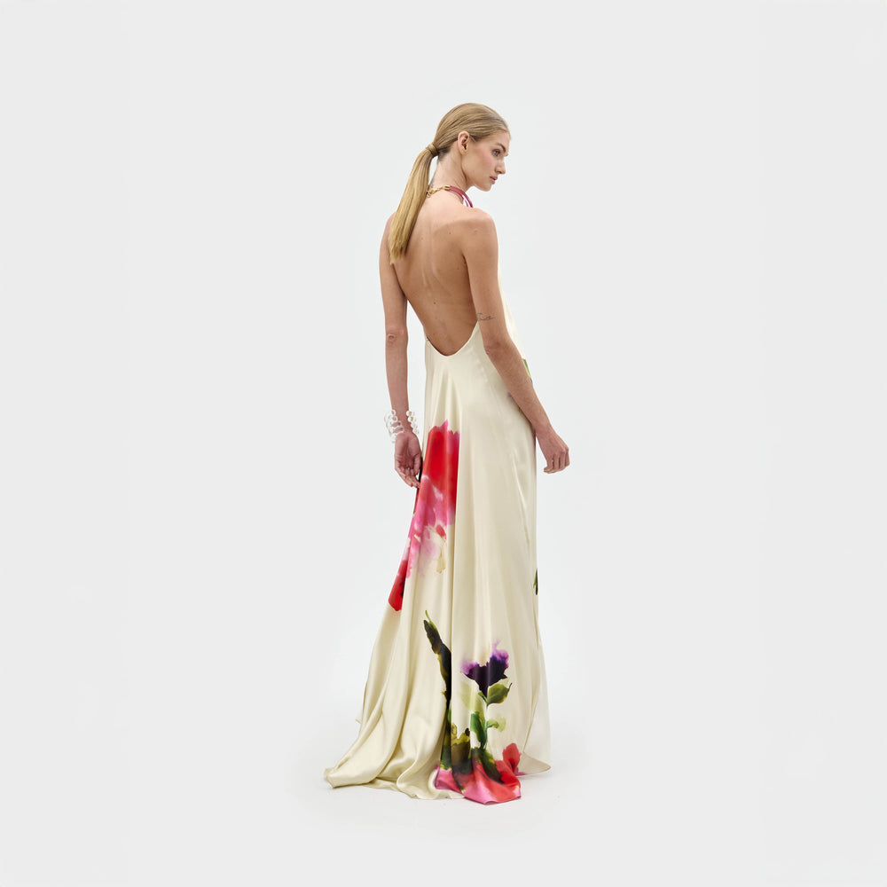A person with long blond hair models the Jesse Dress by SVT DESIGNS, a backless, floor-length maxi with a cream base and bold floral prints, standing against a plain white background and looking over their left shoulder.