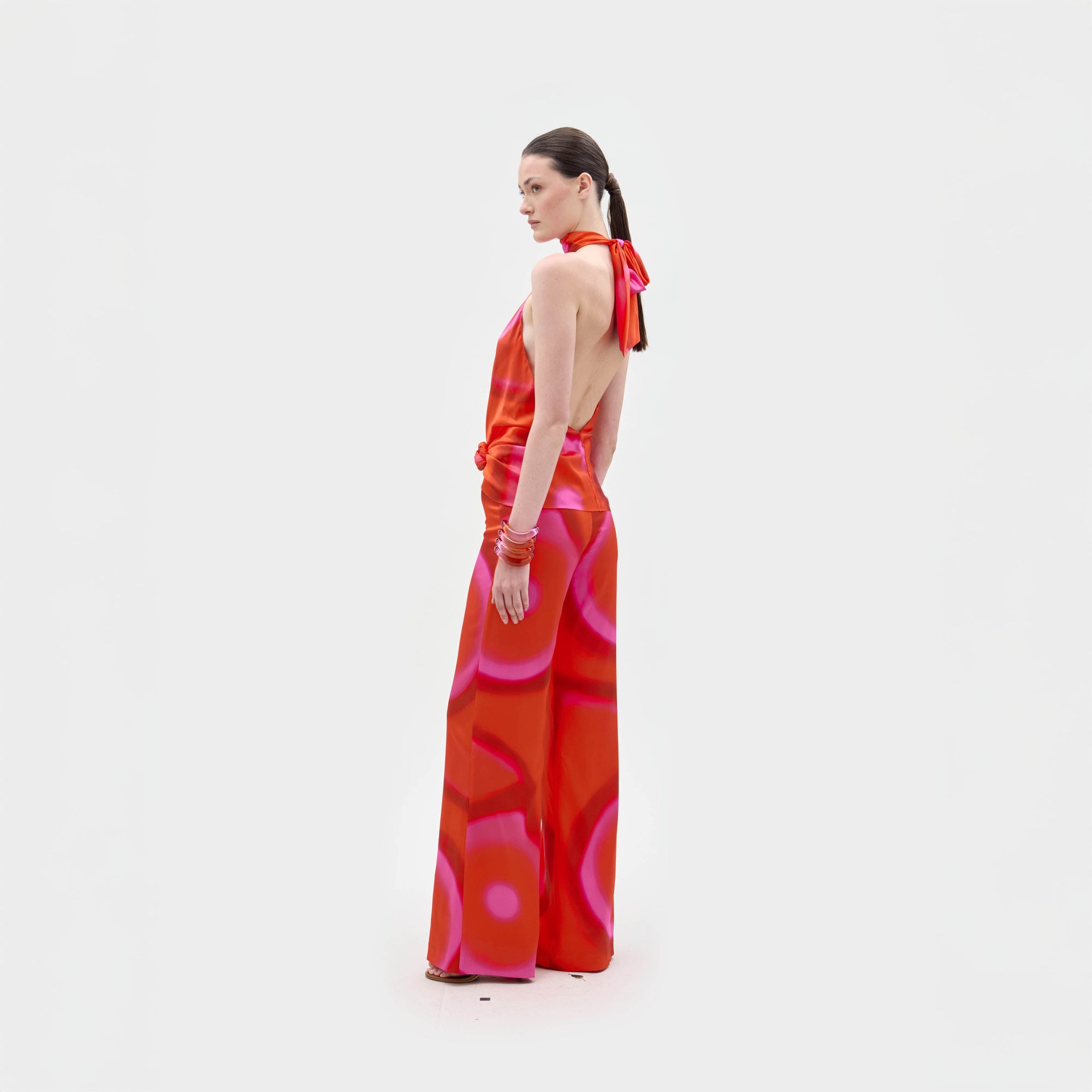 A person stands against a plain white background wearing the Andie Pant by SVT DESIGNS, featuring a bold pink circular pattern on luxurious Italian fabric. They look over their shoulder, showcasing the standout design.