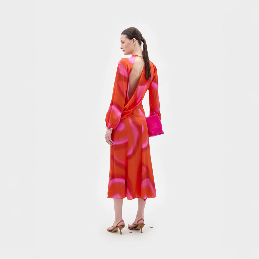 A woman stands facing away against a white background, wearing the Muna Dress by SVT DESIGNS—an orange and pink patterned, long-sleeved open-back midi crafted from Italian fabric—paired with tan heeled sandals and a bright pink clutch.