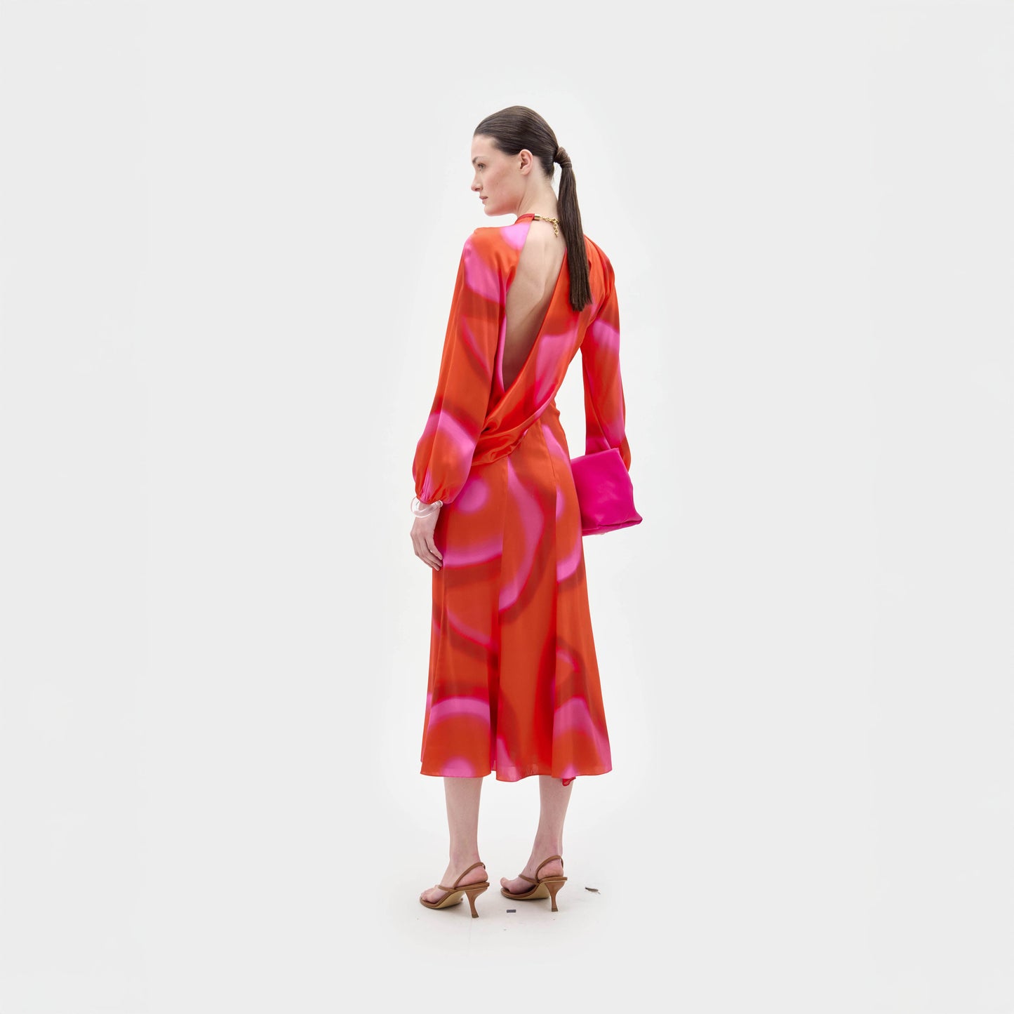 A woman stands facing away against a white background, wearing the Muna Dress by SVT DESIGNS—an orange and pink patterned, long-sleeved open-back midi crafted from Italian fabric—paired with tan heeled sandals and a bright pink clutch.