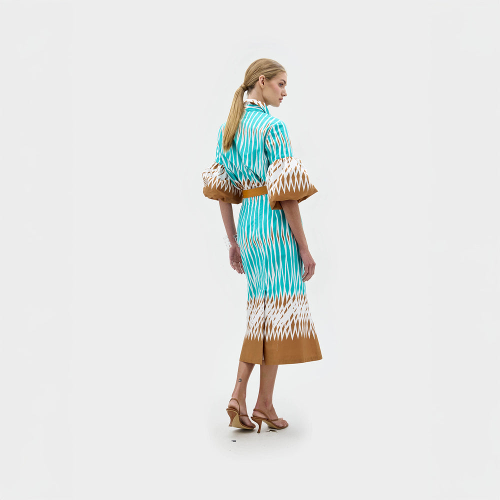 A woman with long blonde hair stands in profile against a plain white background, wearing the Atira Skirt by SVT DESIGNS—a turquoise high-waisted skirt with white and brown patterns, wide sleeves, and brown heeled sandals.