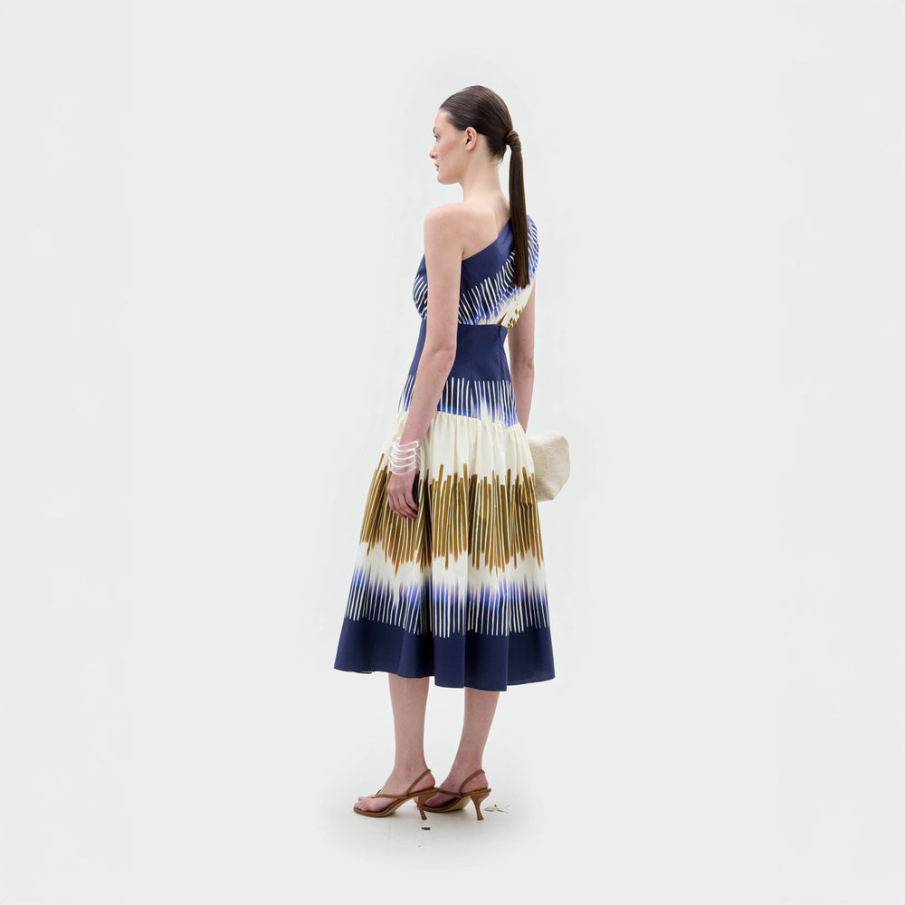 A woman stands against a white background wearing the SVT DESIGNS Catalina Dress—a one-shoulder, sleeveless blue and white midi dress with yellow accents and an A-line silhouette—paired with heeled sandals and a light bag, her hair pulled back.