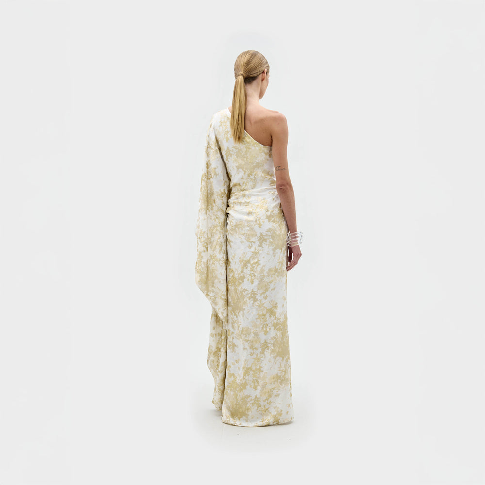 A person with light skin and blonde hair in a low ponytail faces away, modeling the SVT DESIGNS Afrodita Dress—a white and pale yellow floral one-shoulder atelier piece—against a plain white background.