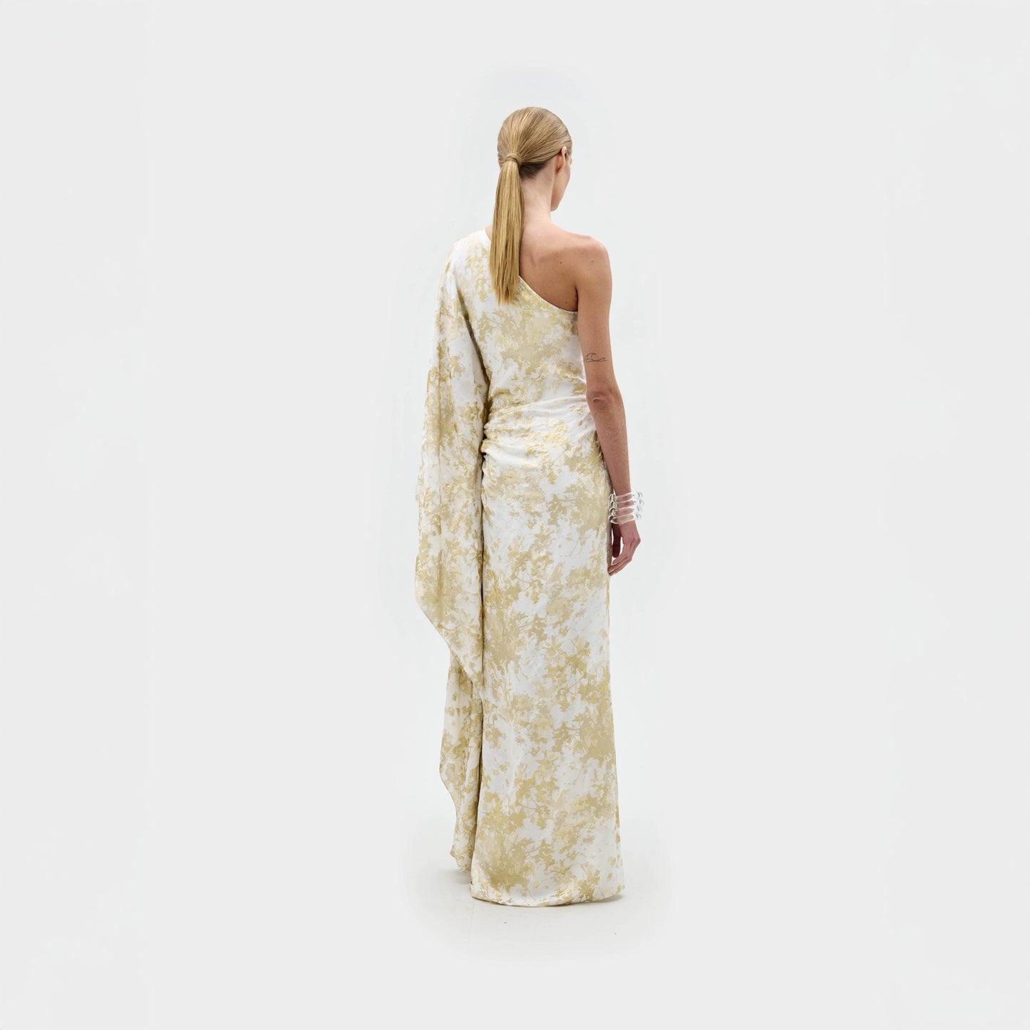 A person with light skin and blonde hair in a low ponytail faces away, modeling the SVT DESIGNS Afrodita Dress—a white and pale yellow floral one-shoulder atelier piece—against a plain white background.