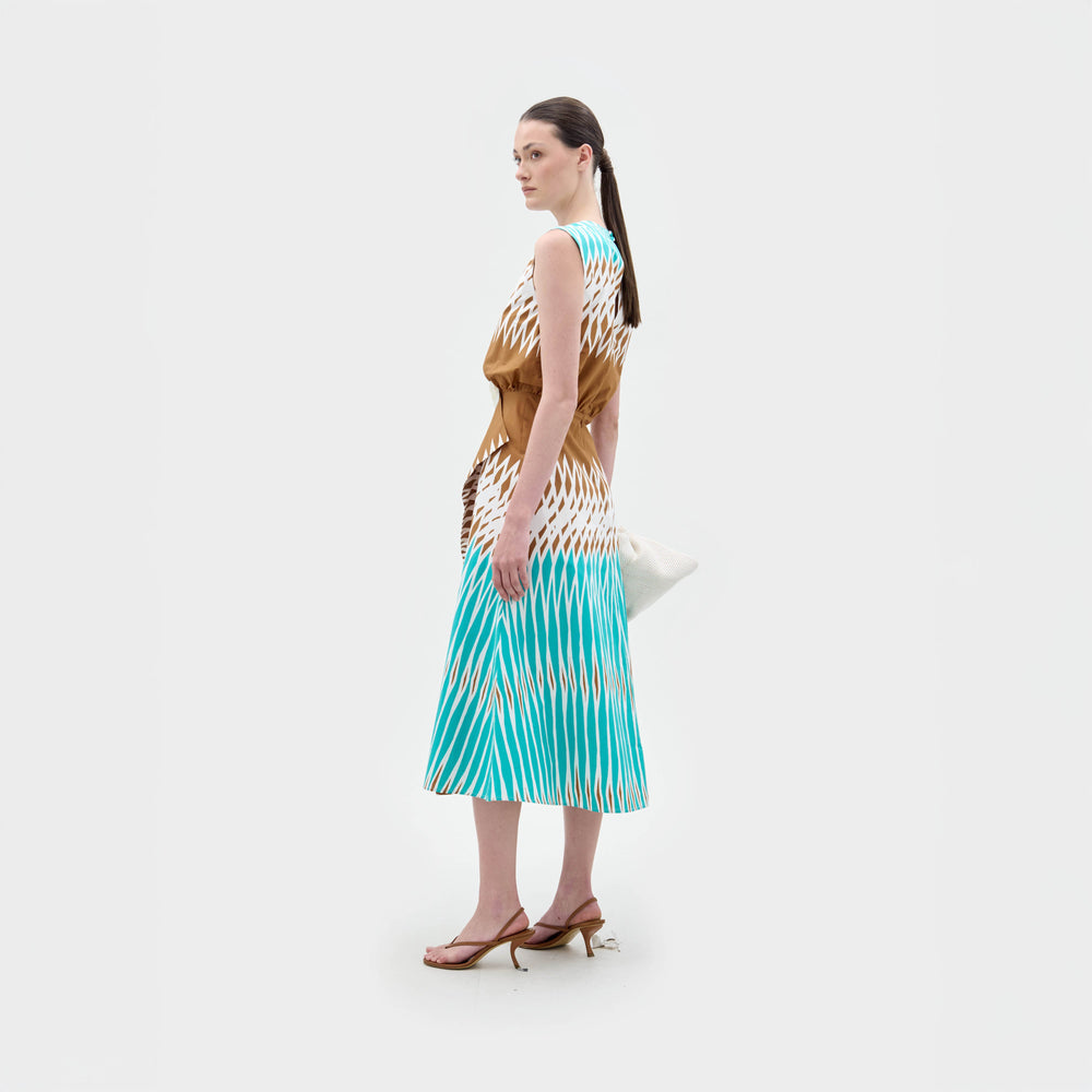 A woman stands sideways against a white background wearing the sleeveless Kendall Dress by SVT DESIGNS, crafted from brown, white, and blue Italian fabric. She accessorizes with a white clutch and brown heeled sandals.