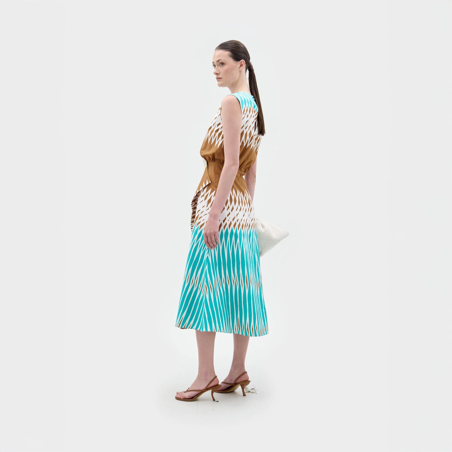A woman stands sideways against a white background wearing the sleeveless Kendall Dress by SVT DESIGNS, crafted from brown, white, and blue Italian fabric. She accessorizes with a white clutch and brown heeled sandals.