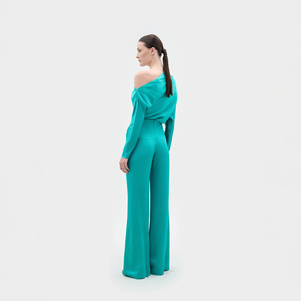 A woman with long dark hair in a ponytail faces away, wearing the SVT DESIGNS Palermo Pant and an off-the-shoulder turquoise blouse against a plain white background.