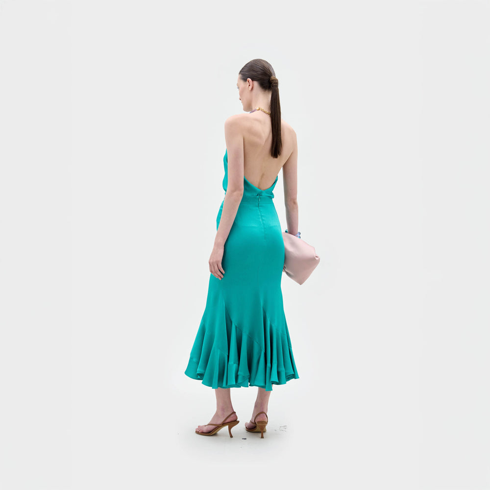 A woman with straight brown hair in a low ponytail stands facing away, wearing the Marlene Skirt by SVT DESIGNS—an A-line teal midi skirt with flared hem—paired with tan heeled sandals, holding a pale pink clutch on a plain white background.