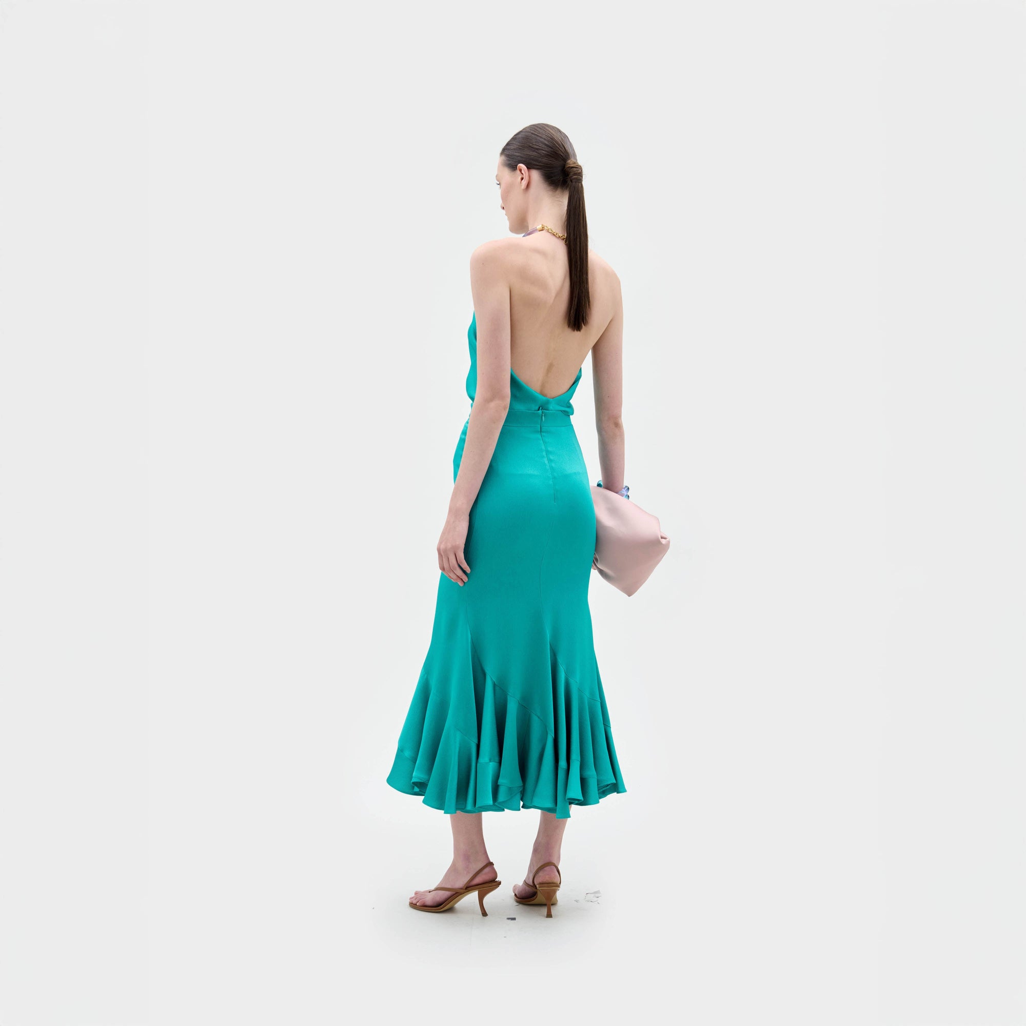 A woman with straight brown hair in a low ponytail stands facing away, wearing the Marlene Skirt by SVT DESIGNS—an A-line teal midi skirt with flared hem—paired with tan heeled sandals, holding a pale pink clutch on a plain white background.