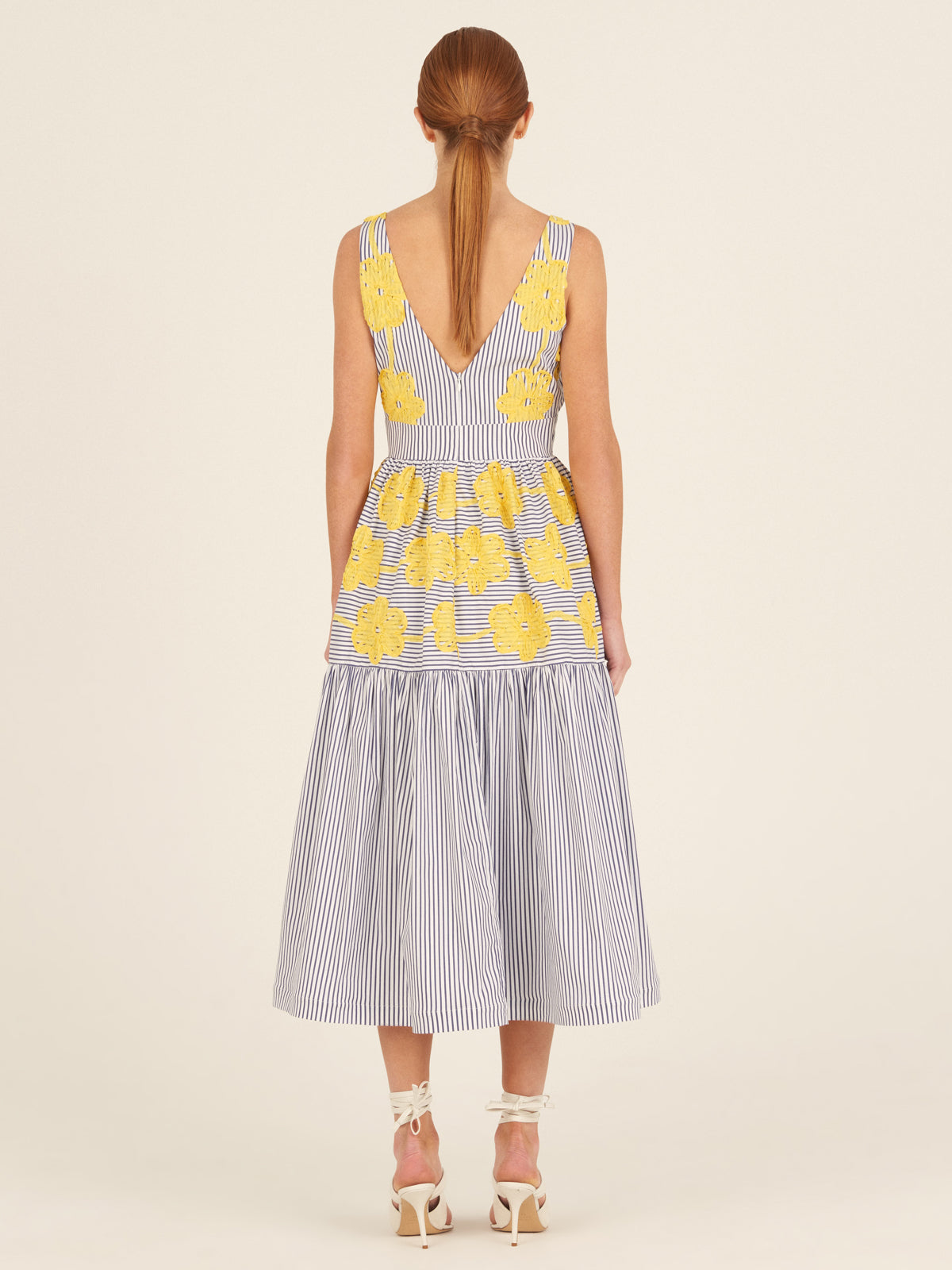 The Eugene Dress, adorned with yellow floral embroidery, showcases a sleeveless design with blue and white vertical stripes, a V-neckline, and a long-tiered skirt.