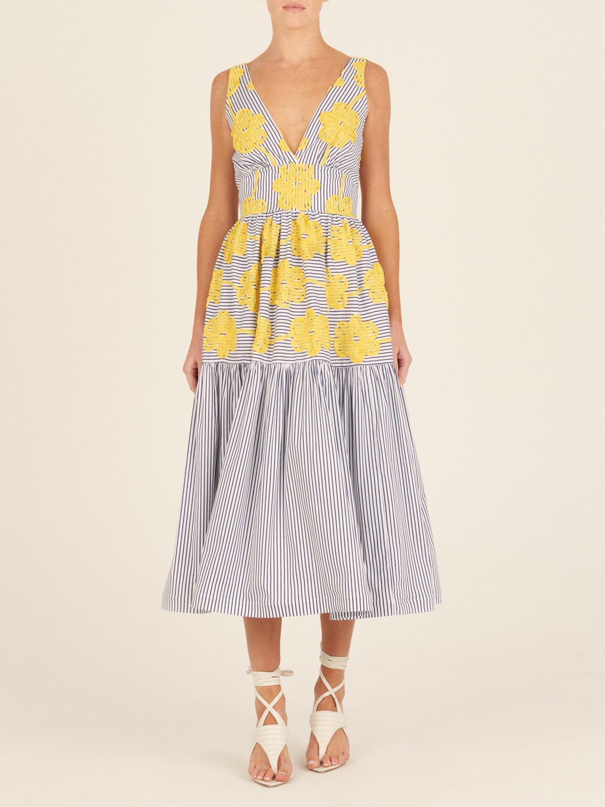 The Eugene Dress, adorned with yellow floral embroidery, showcases a sleeveless design with blue and white vertical stripes, a V-neckline, and a long-tiered skirt.