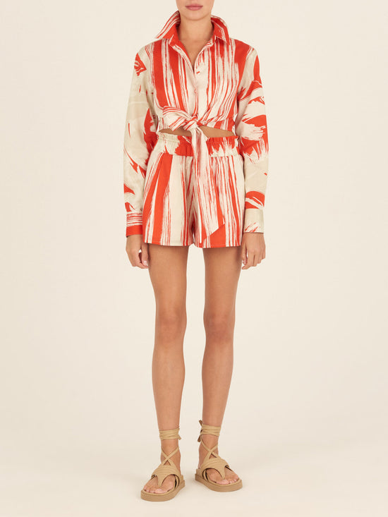 The Gisela Blouse Coral Red Palm Print by Silvia Tcherassi showcases a long-sleeve, button-up design with coral red palm print fabric, featuring abstract stripes and a stylish collar with a tied knot at the waist.