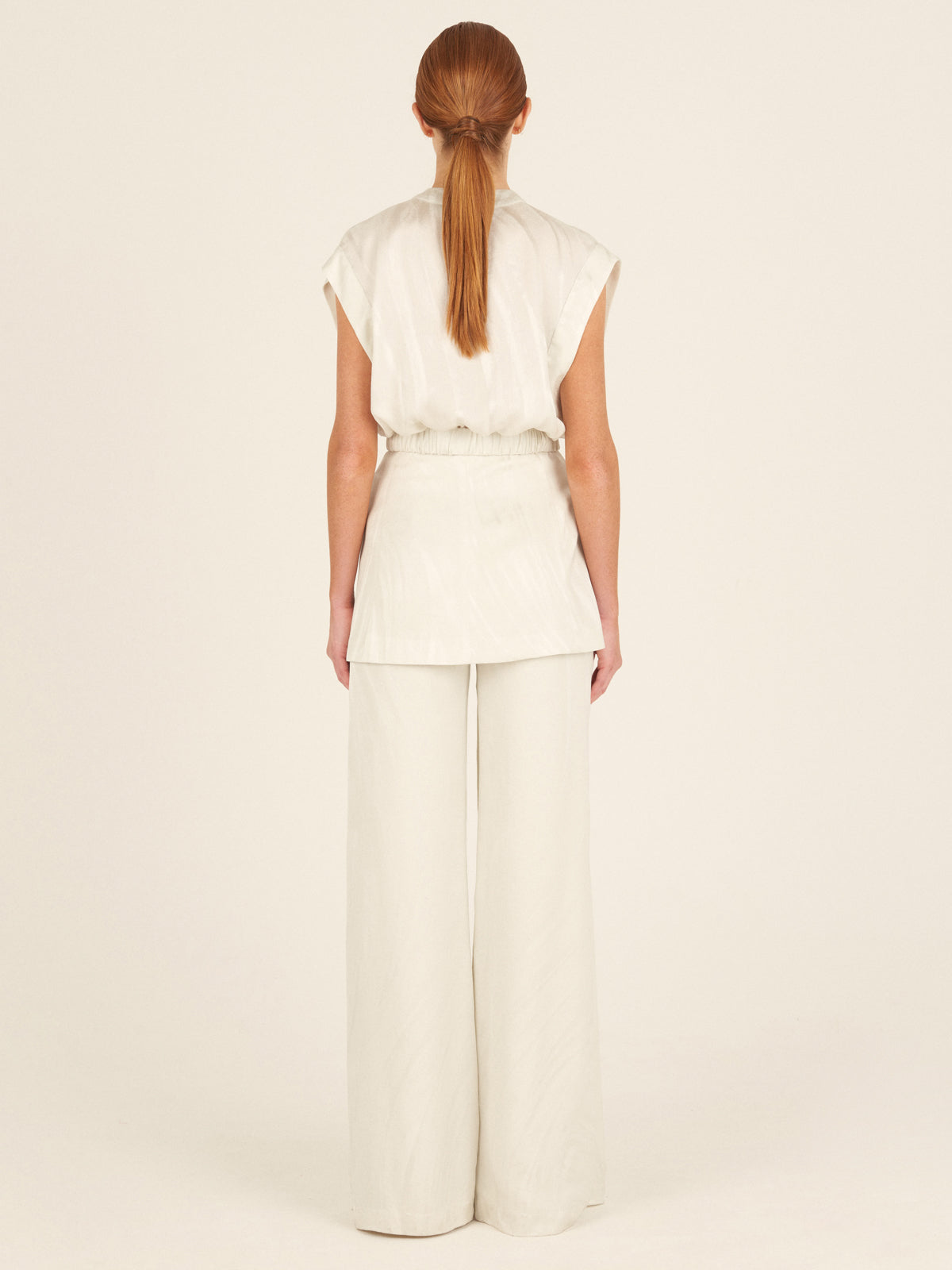 A person with long hair in a ponytail stands elegantly with their back to the camera, wearing the modern Keith Vest Moonstone by Silvia Tcherassi, featuring a sleeveless design and wide-leg trousers in moonstone jacquard fabric, against a plain neutral backdrop.