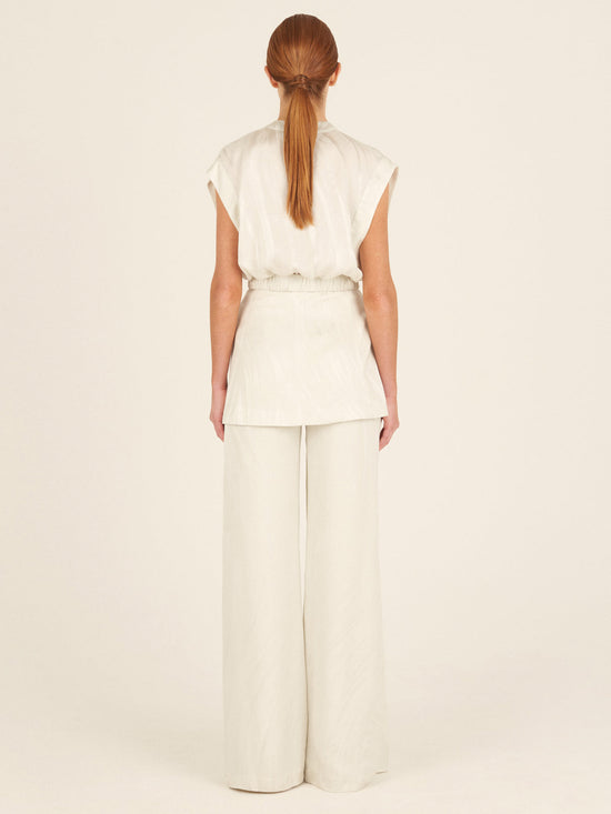 A person with long hair in a ponytail stands elegantly with their back to the camera, wearing the modern Keith Vest Moonstone by Silvia Tcherassi, featuring a sleeveless design and wide-leg trousers in moonstone jacquard fabric, against a plain neutral backdrop.