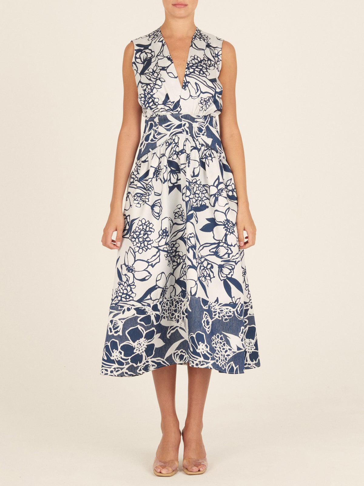 The Nayla Dress Navy Whimsical Garden is a sleeveless white dress with a deep V-neck and a flared skirt, boasting a blue floral print and a charming retro silhouette.