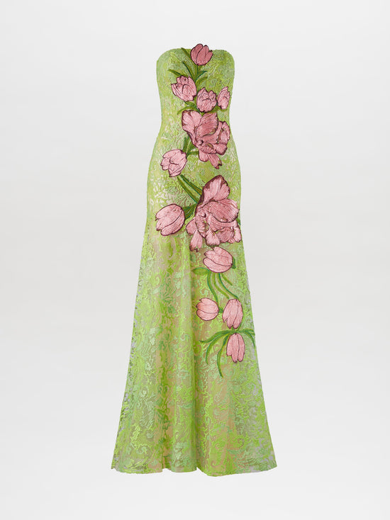 A woman stands wearing the Tulip Dress Rose Tulips, a strapless, green lace dress adorned with large pink floral designs, looking to her left against a plain light background. Pre-order now for exclusive access; shipping by February 1st, 2025.