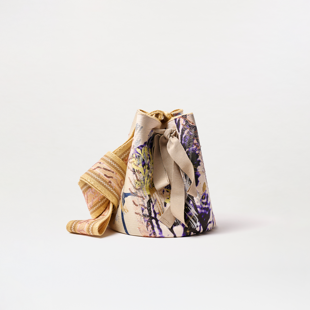 The Mochila Laaput Big is a large fabric bucket bag with a colorful abstract pattern, wide woven shoulder strap, and decorative front bow, displayed on a plain white background.