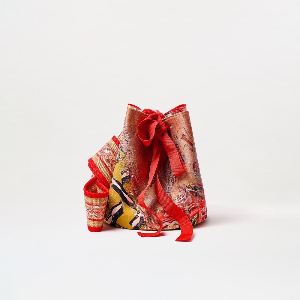 The Mochila Laaput Big is a large red bucket bag with an abstract red, yellow, and orange pattern, a decorative bow, and a wide woven red and cream strap, shown on a plain white background.