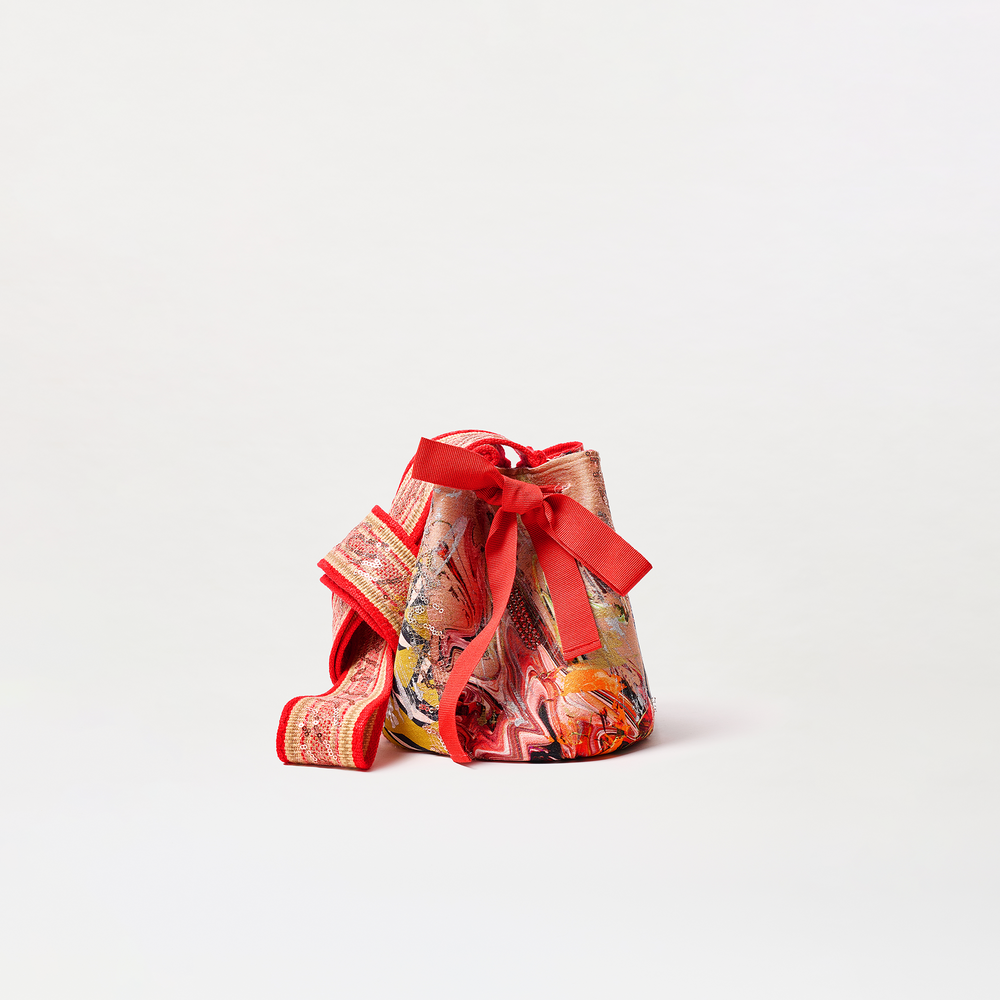 The Mochila Laaput Small is a vibrant Colombian-made handbag featuring a marble-like red, orange, and pink design, a wide red strap, and a large decorative bow, displayed on a plain white background.