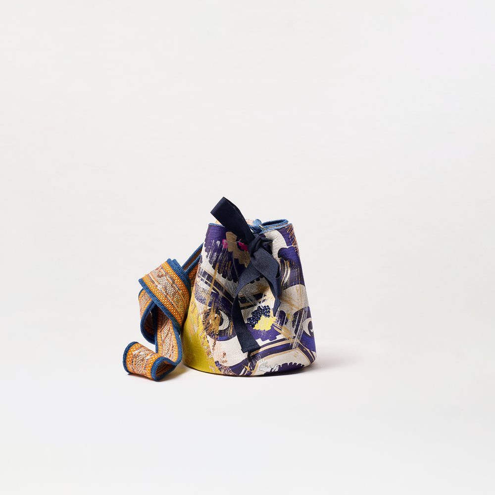 The Mochila Laaput Small is a colorful patterned bucket bag with a wide textured strap and decorative bow, featuring blue, yellow, orange, and white shades. Made in Colombia, it's shown on a plain white background.