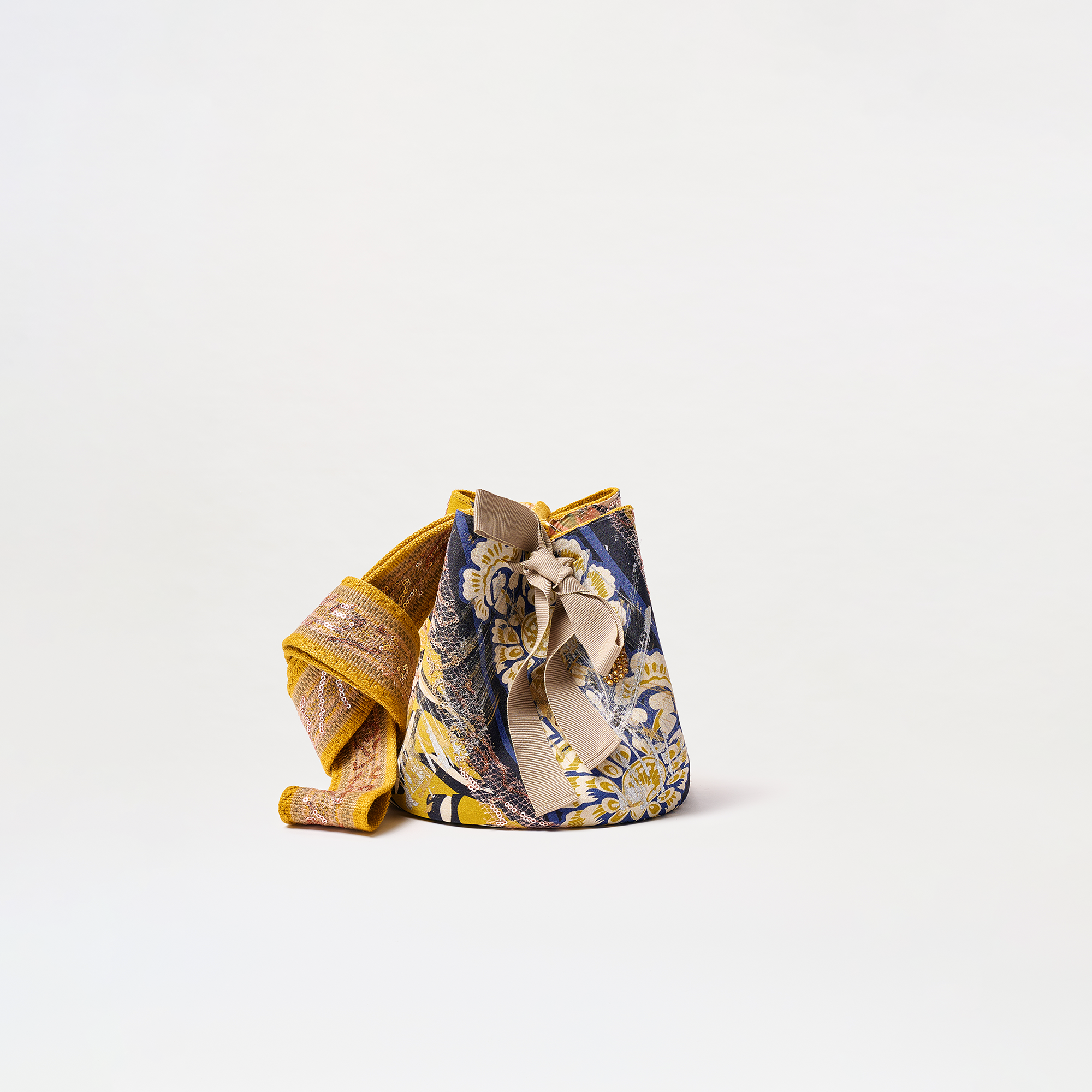 The Mochila Laaput Small is a Colombian-made fabric bag featuring an intricate yellow and blue floral design, a front bow accent, and a woven yellow strap, all set against a plain white background.