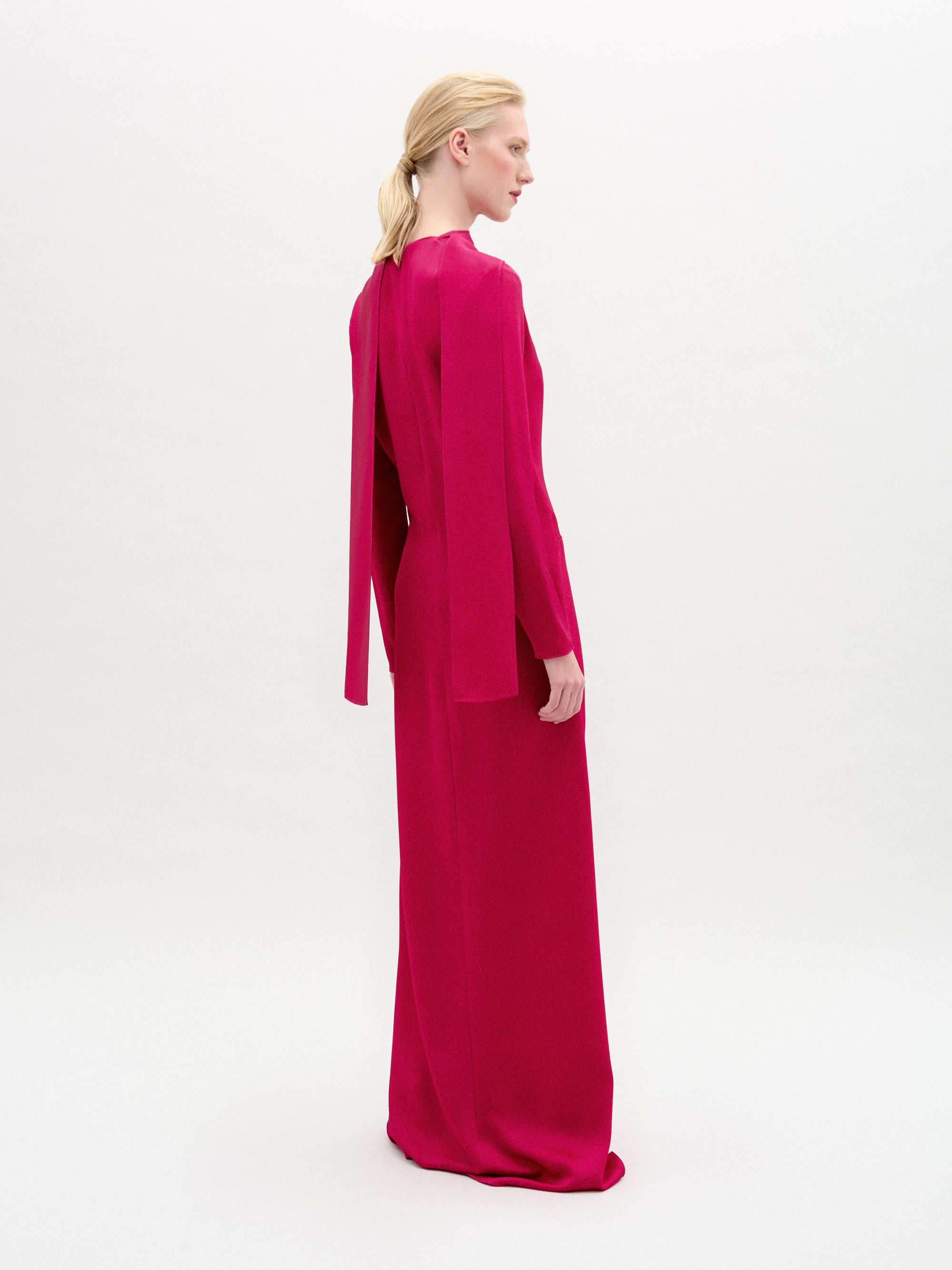 A woman poses against a plain background, slightly turned to the side. She wears the Sue Dress by SVT DESIGNS, crafted from luxurious Italian fabric in vibrant pink. It features a high neckline and long sleeves, with her blonde hair in a low ponytail accentuating the ensemble.