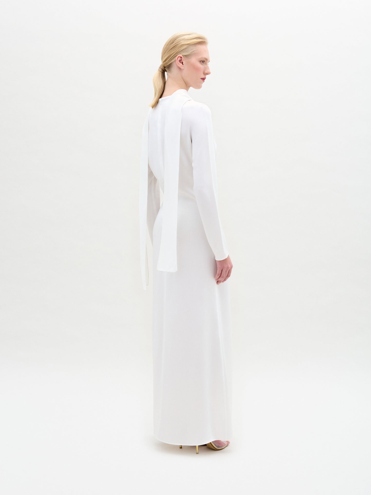 Standing in profile, a person showcases the SVT DESIGNS Sue Dress, a satin maxi with a cape overlay that drapes elegantly from the shoulders. Their hair is styled back against a plain white backdrop.
.