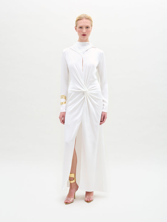 A person poses against a plain background in the Sue Dress by SVT DESIGNS, a satin maxi dress with knot detail and an open front slit. The ensemble includes long sleeves, enhanced by gold arm bands and matching sandals.