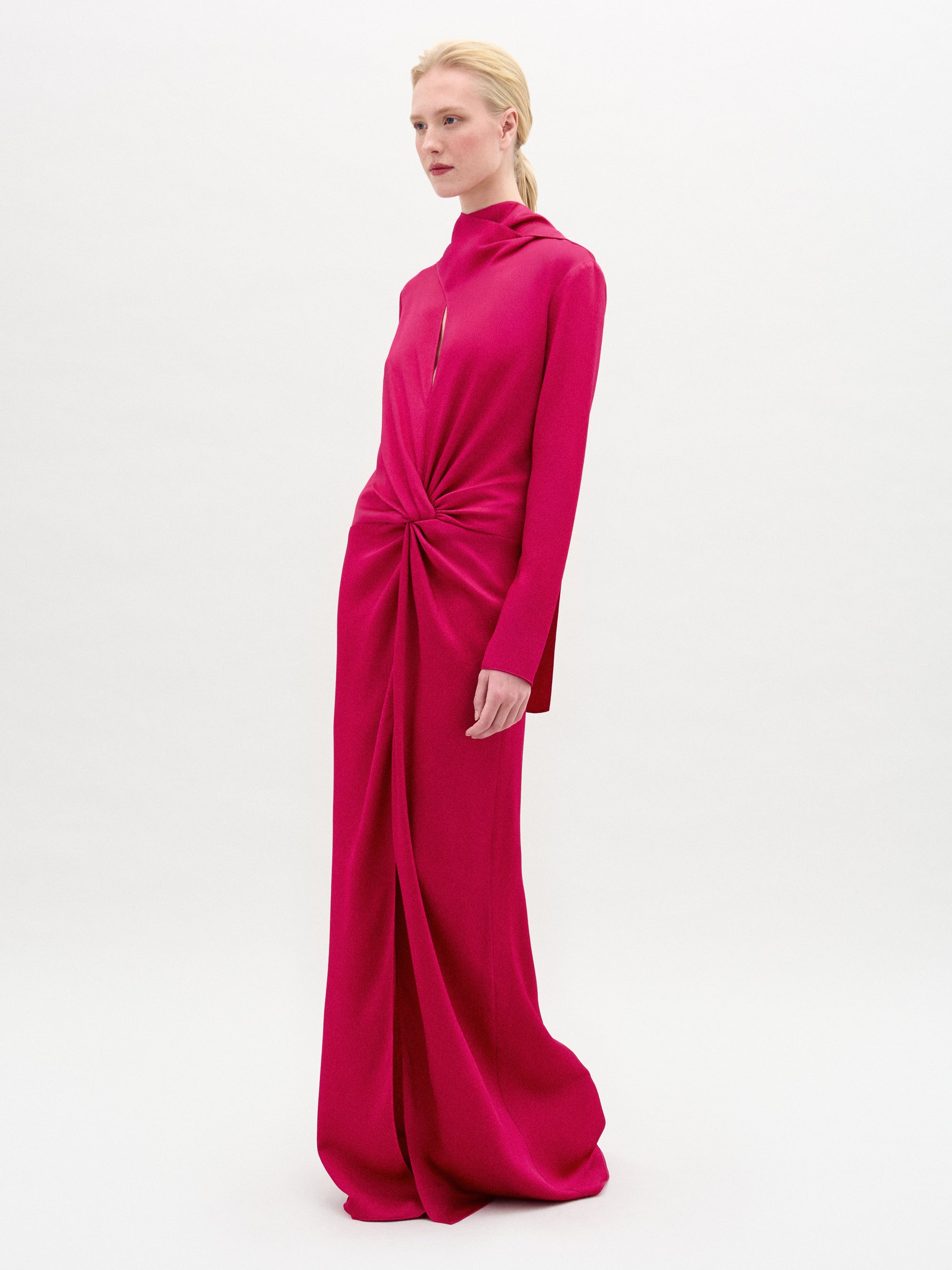 A person stands against a plain white background wearing the Sue Dress by SVT DESIGNS—a long-sleeved, bright pink maxi dress crafted from luxurious Italian fabric, featuring a high neckline and a gathered knot detail at the waist.