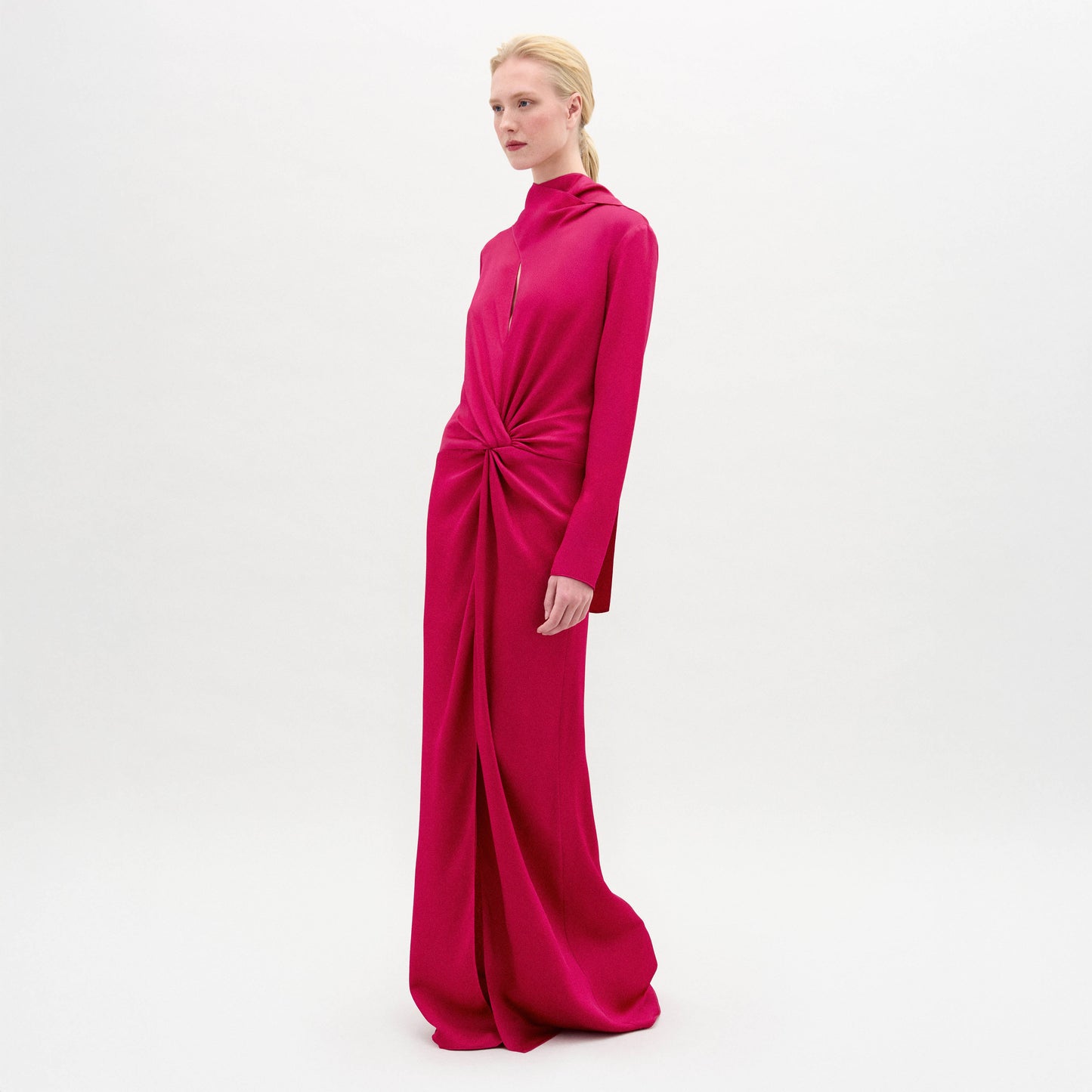 A woman poses in the Sue Dress, a vibrant pink, long-sleeved maxi made from luxurious Italian fabric with a gathered knot at the waist, set against a plain white background.