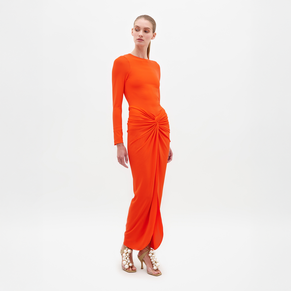 A person wearing the Sydelle Dress Tangerine, a long-sleeved orange dress featuring a knotted detail in the front, stands looking to the side. They are also donning sandals embellished with pearl-like beads. This elegant outfit will be available for pre-order and is anticipated to ship by 2025.