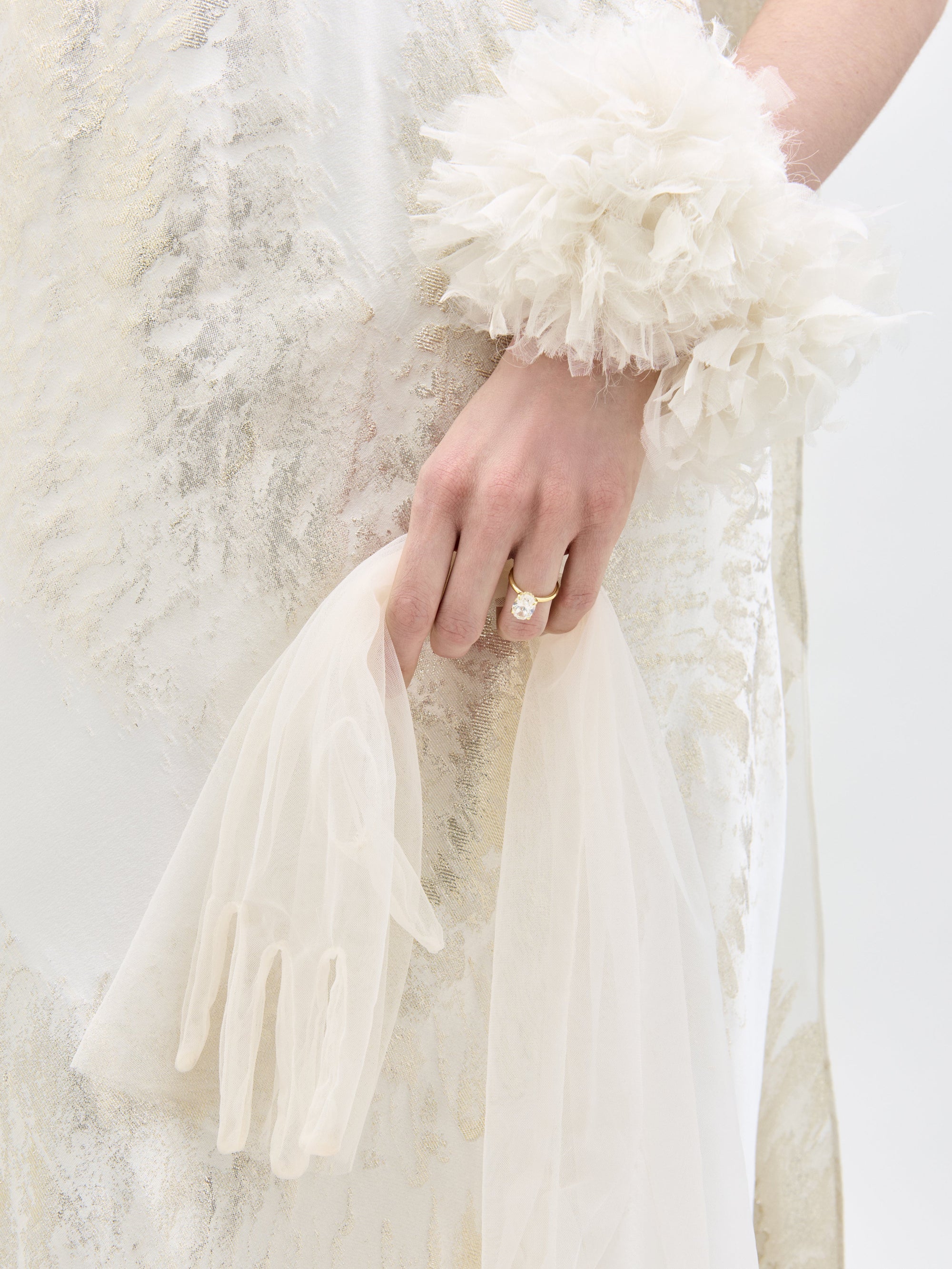 A person holds a sheer, white glove while wearing the Sabina Dress by SVT DESIGNS. They also sport a floral-textured wrist accessory, set against a plain, light-colored background.