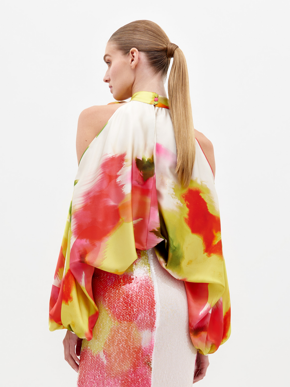 A woman faces away, wearing the SVT DESIGNS Safira Blouse, a halter neck top made from Italian fabric with vibrant red, yellow, and pink abstract patterns. Her hair is in a sleek ponytail and she pairs it with a shimmery pink and white skirt.